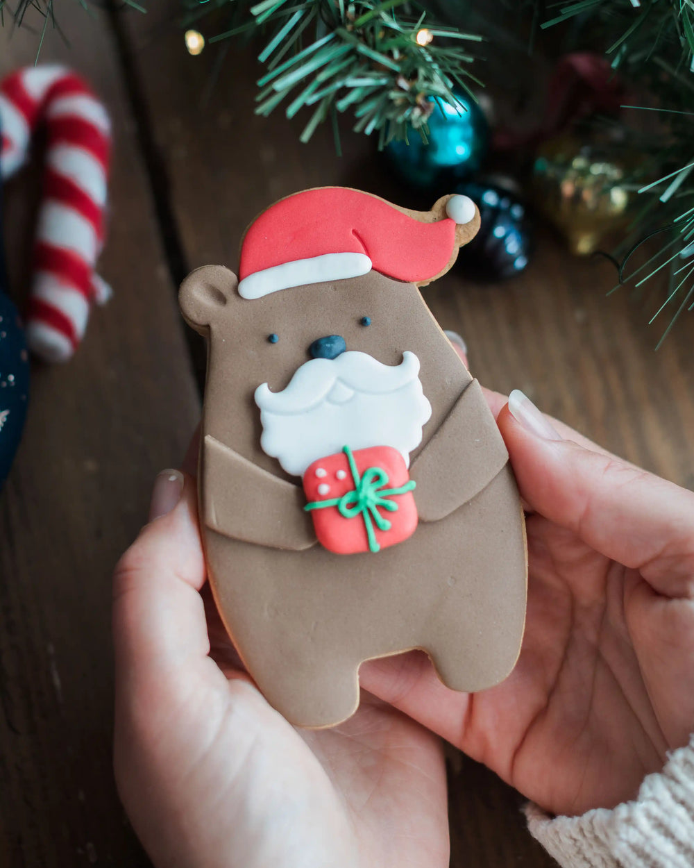 Honeywell Bakes Santa Paws Biscuit Bear - Hand holding a bear-shaped cookie with Santa hat and gift, Christmas decorations in the background