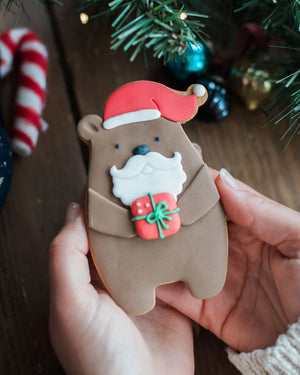 Honeywell Bakes Santa Paws Biscuit Bear - Hand holding a bear-shaped cookie with Santa hat and gift, Christmas decorations in the background