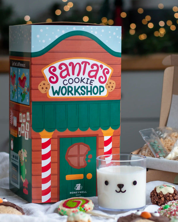 Honeywell Bakes Santa’s Cookie Workshop - 12 Days Christmas Baking Advent - A Honeywell Bakes 'Santa's Cookie Workshop' gift box styled on a table with a glass of milk featuring a bear face and holiday cookies in the foreground, set against a warm, blurred background with festive lights.
