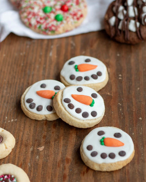Honeywell Bakes Santa’s Cookie Workshop - 12 Days Christmas Baking Advent - Snowman sugar cookies decorated with white icing, chocolate chip eyes and arms, and a small orange carrot nose with green leaf accents, arranged on a wooden surface.