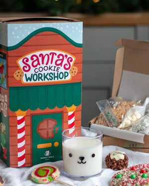 Honeywell Bakes Santa’s Cookie Workshop - 12 Days Christmas Baking Advent - A Honeywell Bakes 'Santa's Cookie Workshop' gift box is shown next to an open box containing assorted cookies, with a glass of milk and decorated cookies in the foreground on a tabletop.