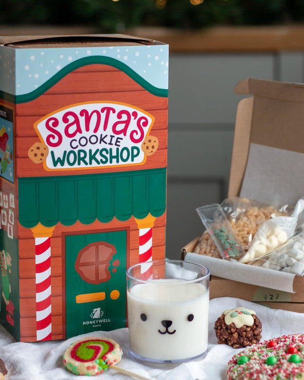 Honeywell Bakes Santa’s Cookie Workshop - 12 Days Christmas Baking Advent - A Honeywell Bakes 'Santa's Cookie Workshop' gift box is shown next to an open box containing assorted cookies, with a glass of milk and decorated cookies in the foreground on a tabletop.
