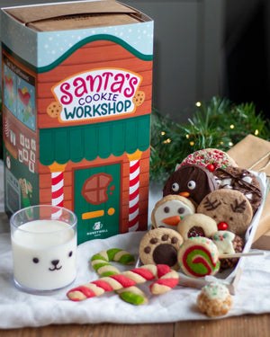 Honeywell Bakes Santa’s Cookie Workshop - 12 Days Christmas Baking Advent - Santa's Cookie Workshop gift box by Honeywell Bakes alongside assorted holiday cookies, candy canes, and a glass of milk on a tabletop with pine branches in the background.