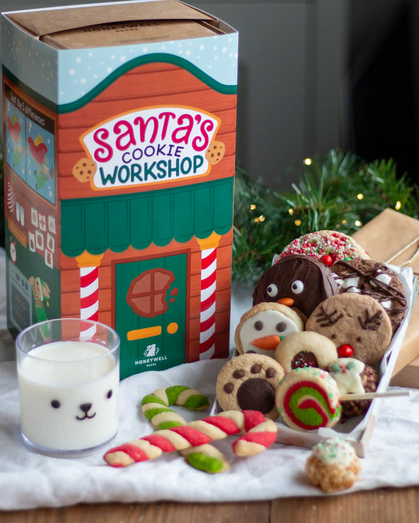 Honeywell Bakes Santa’s Cookie Workshop - 12 Days Christmas Baking Advent - Santa's Cookie Workshop gift box by Honeywell Bakes alongside assorted holiday cookies, candy canes, and a glass of milk on a tabletop with pine branches in the background.