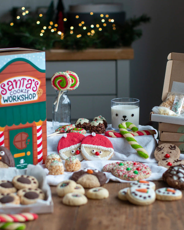 Honeywell Bakes Santa’s Cookie Workshop - 12 Days Christmas Baking Advent - A festive holiday cookie assortment displayed on a wooden table, featuring Santa-themed cookies, candy canes, lollipops, and a glass of milk, alongside a 'Santa's Cookie Workshop' box and a gift box in the background with warm holiday lights.