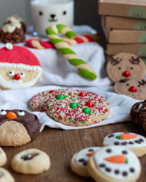 Honeywell Bakes Santa’s Cookie Workshop - 12 Days Christmas Baking Advent - A styled holiday cookie display on a wooden table with a white cloth. The main featured product is a round sugar cookie topped with red and green candy and white sprinkles, surrounded by other decorated Christmas cookies including a Santa face, snowman, penguin, and reindeer, along with candy canes and gift boxes.