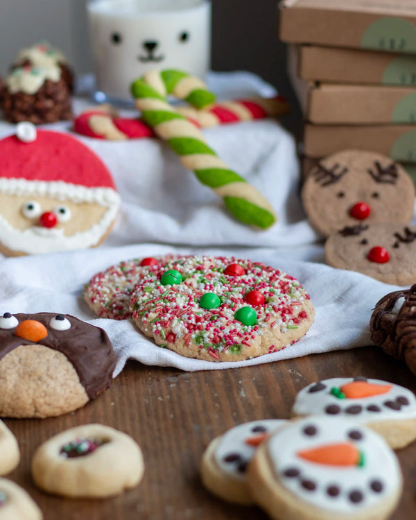 Honeywell Bakes Santa’s Cookie Workshop - 12 Days Christmas Baking Advent - A styled holiday cookie display on a wooden table with a white cloth. The main featured product is a round sugar cookie topped with red and green candy and white sprinkles, surrounded by other decorated Christmas cookies including a Santa face, snowman, penguin, and reindeer, along with candy canes and gift boxes.