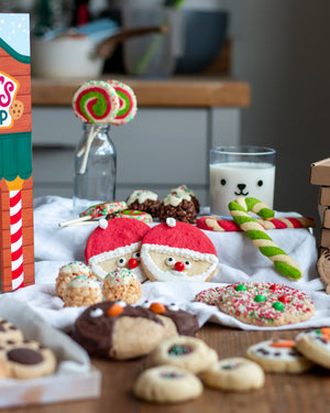 Honeywell Bakes Santa’s Cookie Workshop - 12 Days Christmas Baking Advent - A styled holiday cookie spread on a wooden table, featuring Santa face cookies, candy canes, chocolate-dipped cookies, a lollipop in a glass bottle, a glass of milk with a bear face, and a colourful candy cane box on the left.