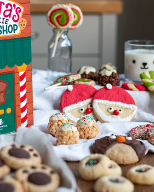 Honeywell Bakes Santa’s Cookie Workshop - 12 Days Christmas Baking Advent - A styled holiday cookie display with Santa face cookies, chocolate chip cookies, rice crispy treats, and a lollipop in a glass bottle, with a box labeled ‘Honeywell Bakes Cookie Shop' visible on the left.