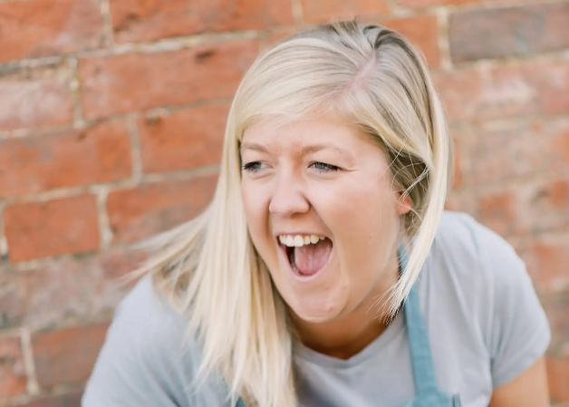 Woman with blonde hair and a blue apron standing in front of a brick wall