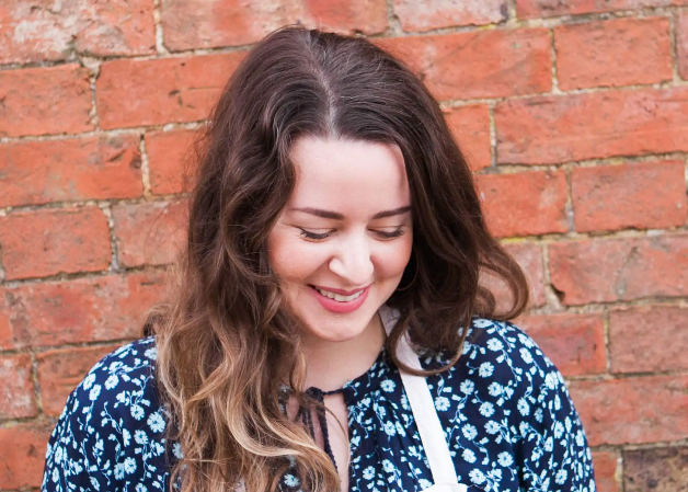 Woman with long brown hair wearing a blue floral blouse against a brick wall.