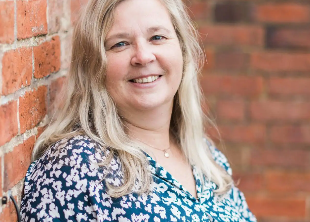 Woman with blonde hair wearing a blue floral blouse standing against a brick wall.