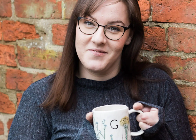 Person holding a mug with a floral design and letter 'G' against a brick wall.