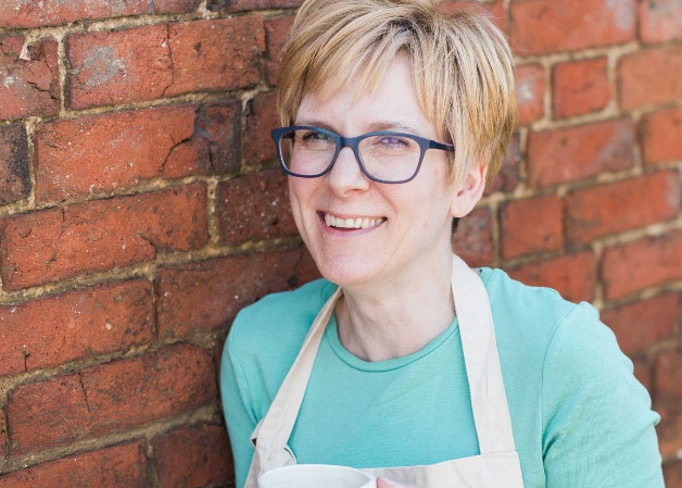 Person wearing glasses and an apron, holding a mug against a brick wall.