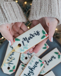 Honeywell Bakes Six Personalised Christmas Tag Biscuits - Hand holding a decorated cookie with 'Maris' written on it, surrounded by other similar cookies.