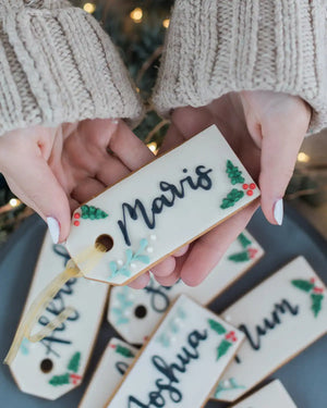 Honeywell Bakes Six Personalised Christmas Tag Biscuits - Hand holding a decorated cookie with 'Maris' written on it, surrounded by other similar cookies.