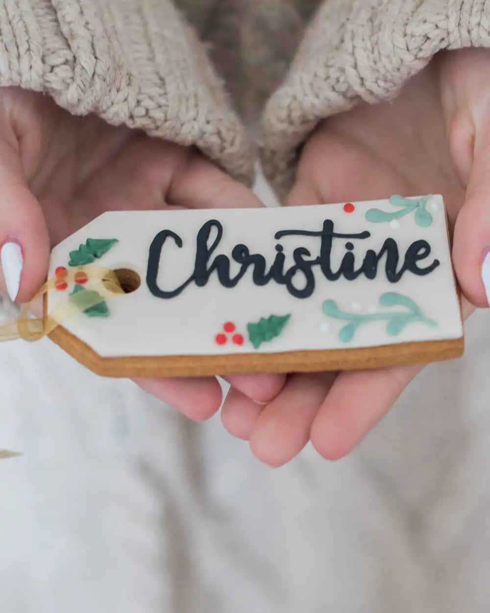 Honeywell Bakes Six Personalised Christmas Tag Biscuits - Hand holding a decorated cookie with 'Maris' written on it, surrounded by other similar cookies.