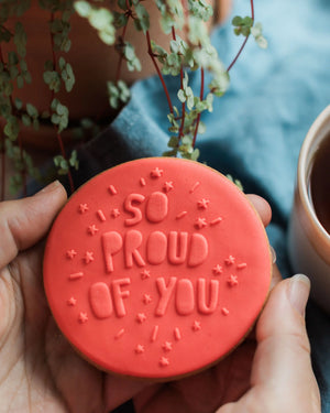 Honeywell Bakes So Proud Of You - Red circular biscuit with 'So Proud of You' text held by a person, with plants and a cup in the background.