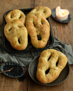 Honeywell Bakes - A golden-brown baked bread fougasse roll with a distinctive ring-like cutout shape, served on a dark plate on a rustic wooden table with a lit candle, more rolls on a tray, and gray linen cloth.