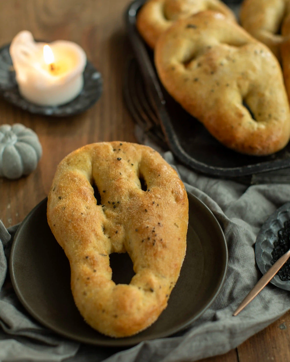 Honeywell Bakes - A golden-brown baked bread fougasse roll with a distinctive ring-like cutout shape, served on a dark plate on a rustic wooden table with a lit candle, more rolls on a tray, and gray linen cloth.