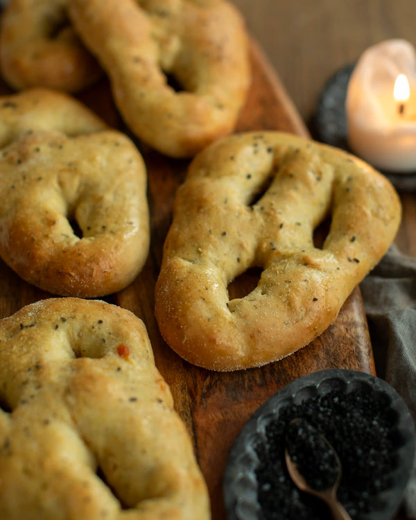 Honeywell Bakes - A golden-brown baked bread fougasse roll with a distinctive ring-like cutout shape, served on a dark plate on a rustic wooden table with a lit candle, more rolls on a tray, and gray linen cloth.