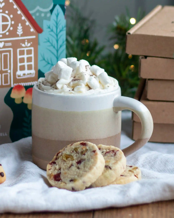 Honeywell Bakes Stoneware Mug - Oatmeal & White - A ceramic mug filled with a creamy beverage topped with whipped cream and marshmallows, styled with two cookies in front and holiday decorations in the background.