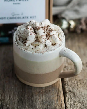 Honeywell Bakes Stoneware Mug - Oatmeal & White - A ceramic mug filled with a hot chocolate drink topped with whipped cream, marshmallows, and cocoa powder, set on a rustic wooden surface with a Honeywell hot chocolate kit box blurred in the background.