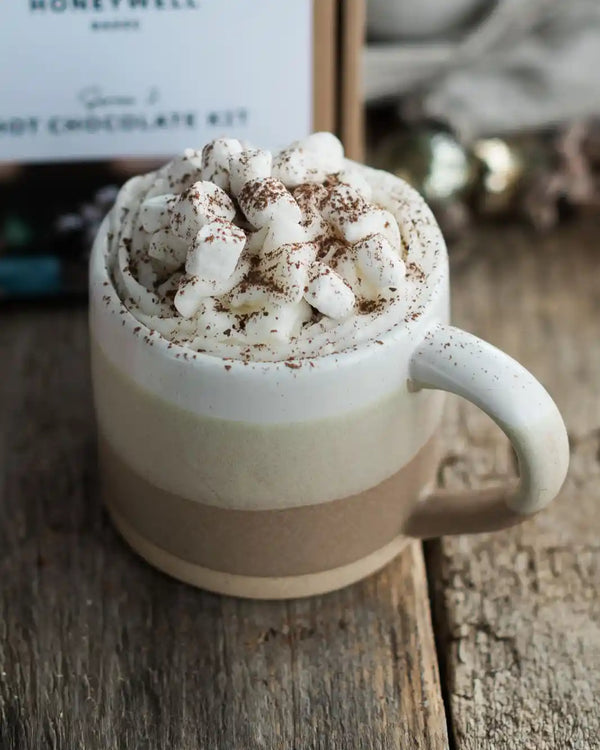 Honeywell Bakes Stoneware Mug - Oatmeal & White - A ceramic mug filled with a hot chocolate drink topped with whipped cream, marshmallows, and cocoa powder, set on a rustic wooden surface with a Honeywell hot chocolate kit box blurred in the background.
