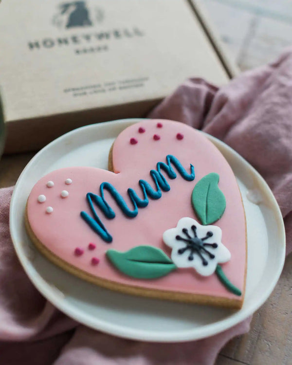 Honeywell Bakes Sweetheart Biscuit – Fully Iced Personalised Heart - Heart-shaped cookie with ‘Mum' text on a plate with a cup of gift box in the background.