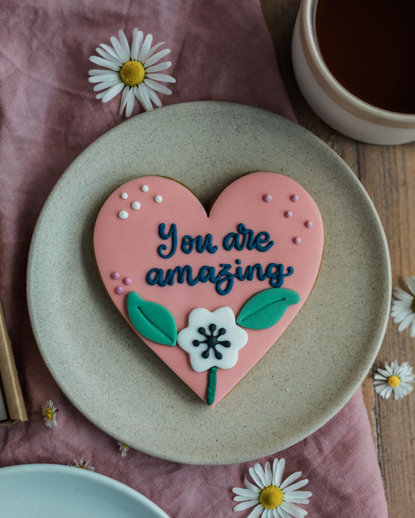 Honeywell Bakes Sweetheart Biscuit – Fully Iced Personalised Heart - Heart-shaped cookie with ‘You are amazing' on a plate with a cup of coffee and daisies.
