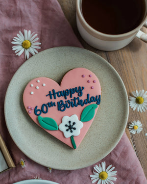 Honeywell Bakes Sweetheart Biscuit – Fully Iced Personalised Heart - Heart-shaped cookie with 'Happy 60th Birthday' on a plate with a cup of coffee and daisies.