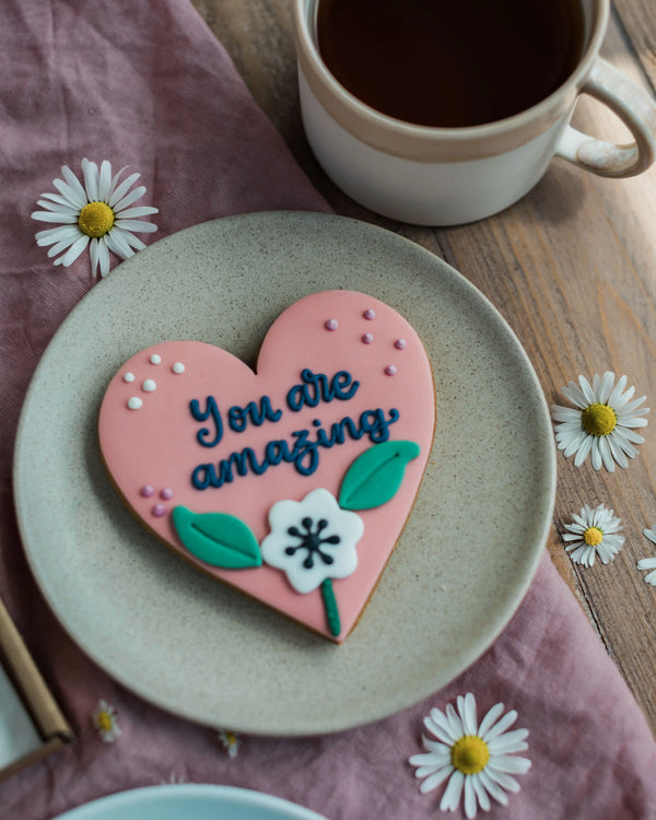 Honeywell Bakes Sweetheart Biscuit – Fully Iced Personalised Heart - Heart-shaped cookie with 'You are amazing' text on a plate with a cup of coffee and daisies.
