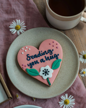 Honeywell Bakes Sweetheart Biscuit – Fully Iced Personalised Heart - Heart-shaped cookie with 'Sending you a hug' text on a plate with a cup of coffee and daisies.