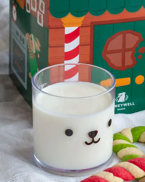 Honeywell Bakes Teddy Tumbler Cup - A clear glass tumbler with a bear face design, filled with white milk, set in front of a colourful Honeywell Bakes holiday box. Candy cane cookies are visible on the right side.