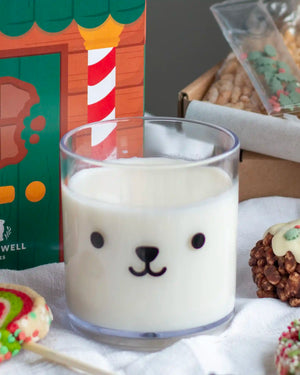 Honeywell Bakes Teddy Tumbler Cup - A clear cylindrical glass filled with milk and a cute black bear face printed on the front, styled on a white cloth with holiday treats and a colourful gift box in the background.