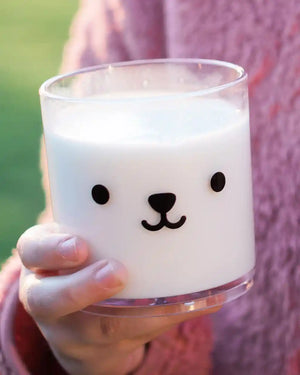 Honeywell Bakes Teddy Tumbler Cup - A hand holding a clear glass candle jar filled with milk, decorated with a cute black bear face. The person holding the cup is wearing a pink knit sweater, and the background is a soft-focus green outdoor setting.