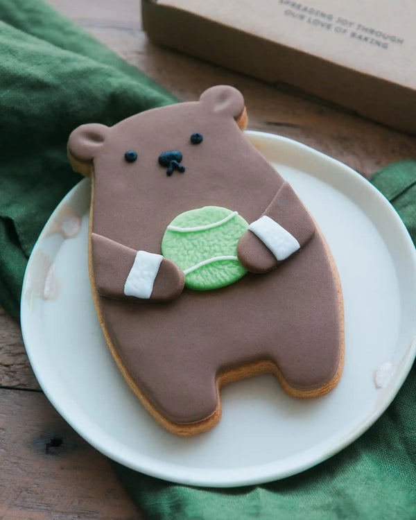 Honeywell Bakes Tennis Biscuit Bear - Bear-shaped cookie with a tennis ball on a white plate, with a green napkin and 'Honey Bakes' box in the background.