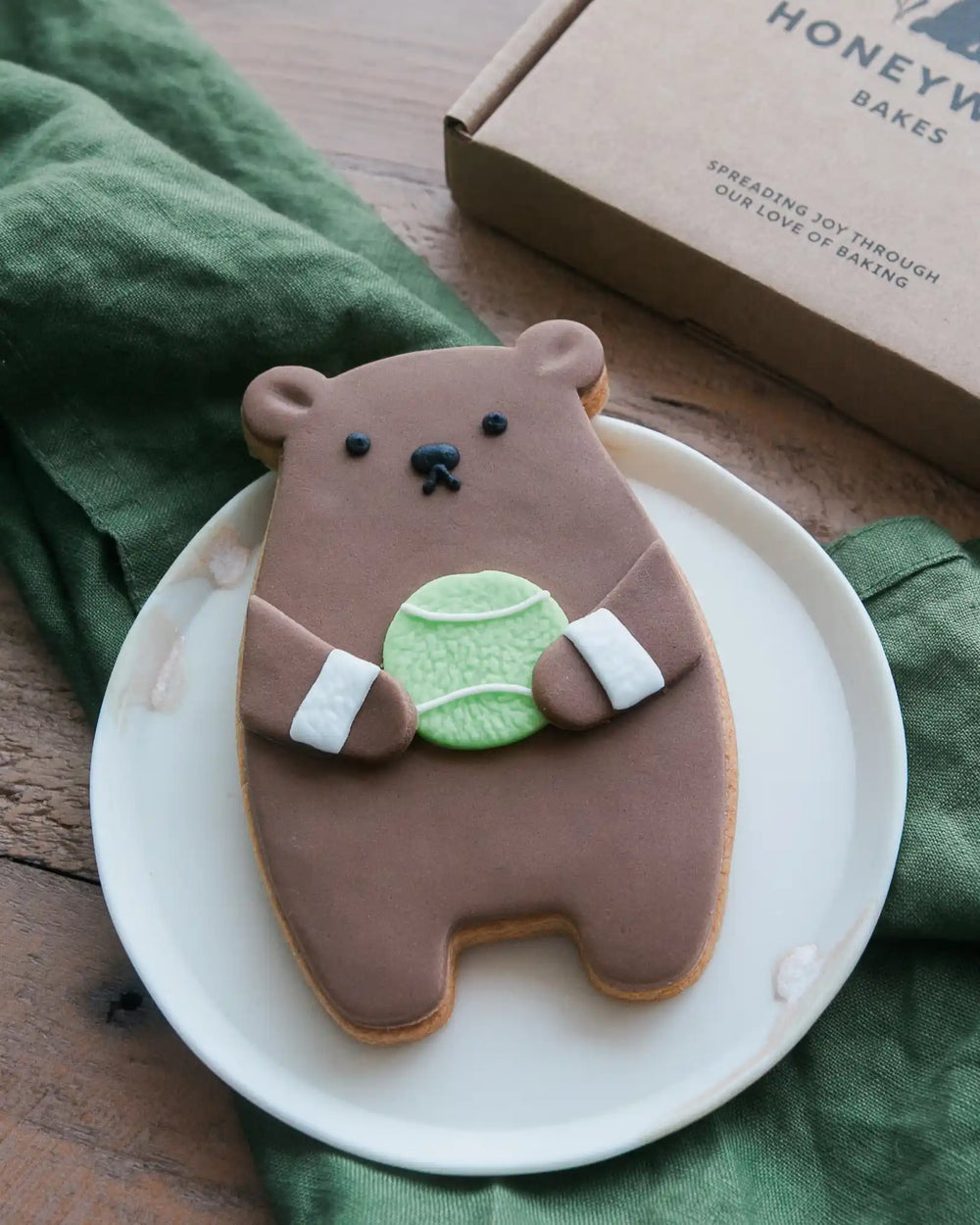 Honeywell Bakes Tennis Biscuit Bear - Bear-shaped cookie with a tennis ball on a white plate, with a green napkin and 'Honey Bakes' box in the background.