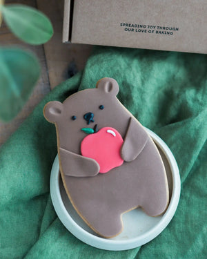 Honeywell Bakes Thank You Teacher Bear Biscuit - Cookie shaped like a bear holding an apple on a green cloth with a box in the background.