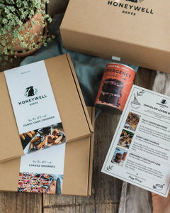 Chocolate Lovers Bundle - Honeywell Bakes gift set arranged on a rustic wooden table, including a kraft gift box, a can of emergency chocolate cake mix, a printed chocolate lover's guide, and a box of loaded brownies.