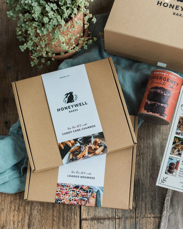Chocolate Lovers Bundle - Honeywell Bakes gift box set on a rustic wooden table, featuring a kraft paper box with a candy cane churros label, another box with a loaded brownie label, an orange emergency chocolate cake can, and a potted green plant.