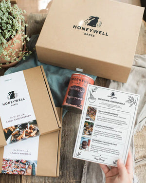 Chocolate Lovers Bundle - Honeywell Bakes gift bundle set displayed on a rustic wooden table, including a large brown kraft box, a smaller box with candy cane churros, a can of emergency chocolate cake mix, and a printed chocolate lovers bundle recipe card held by a hand.
