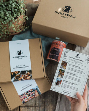 Chocolate Lovers Bundle - Honeywell Bakes chocolate lover's bundle gift set displayed on a rustic wooden table, featuring a brown kraft box with a white paper band, an orange emergency cake tin, a recipe booklet held by a hand, and another box in the background.