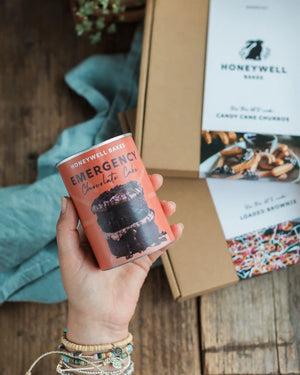 Chocolate Lovers Bundle - A hand holds a Honeywell Bakes Emergency Chocolate Cake tin can, with its cardboard packaging and recipe cards visible in the background on a rustic wooden table.