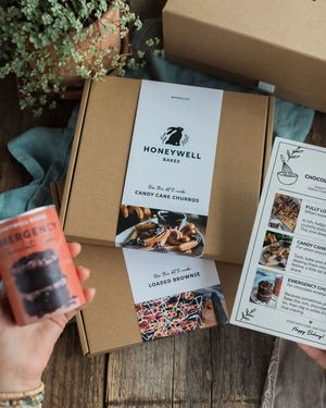 Chocolate Lovers Bundle - A Honeywell Bakes baking kit displayed in a styled flat-lay setting on a rustic wooden table. The kit includes a brown kraft box with a white paper label, a recipe booklet, and a small orange emergency chocolate cake can held by a hand.