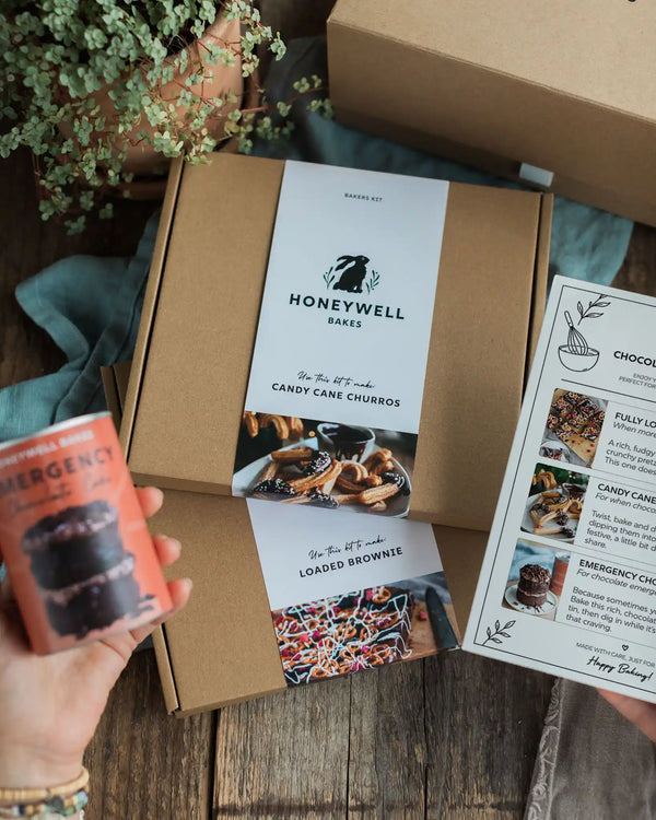 Chocolate Lovers Bundle - A Honeywell Bakes baking kit displayed in a styled flat-lay setting on a rustic wooden table. The kit includes a brown kraft box with a white paper label, a recipe booklet, and a small orange emergency chocolate cake can held by a hand.
