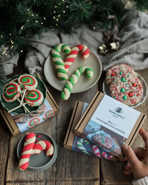 The Christmas Elf Baking Kit Collection - A hand holds a Honeywell House gift box of Christmas eve cookies on a rustic wooden table, surrounded by decorated holiday cookies, candy canes, and pine branches.