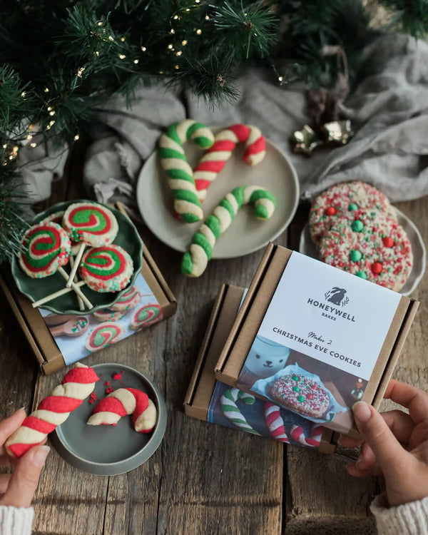 The Christmas Elf Baking Kit Collection - A person's hands hold a Honeywell Nest boxed cookie gift set on a rustic wooden table, surrounded by decorated holiday cookies, candy canes, and pine branches with warm string lights.