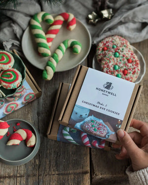 The Christmas Elf Baking Kit Collection - A hand holds a brown cardboard box labeled Honeywell Bakes Christmas Eve Cookies, surrounded by decorated holiday cookies on plates and a rustic wooden table.