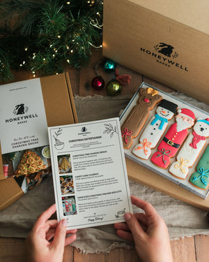 The Christmas Entertainer Bundle by Honeywell Bakes, featuring hand-iced Christmas cracker biscuits, a Christmas tree sharing bread baking kit and chocolate-dipped candy cane churros, presented in festive gift packaging.